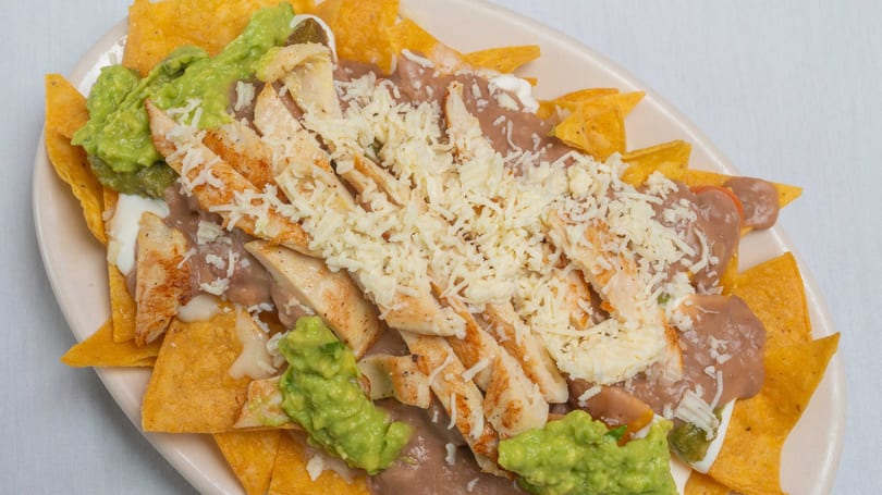 Chicken nachos with refried beans, guacamole, sour cream, and shredded cheese on a white plate.