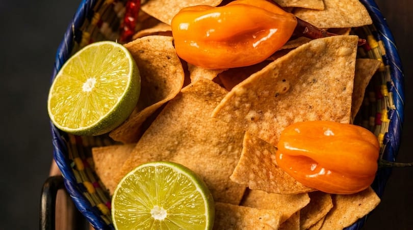 Crispy tortilla chips in a colorful basket, garnished with fresh lime halves and vibrant orange habanero peppers.