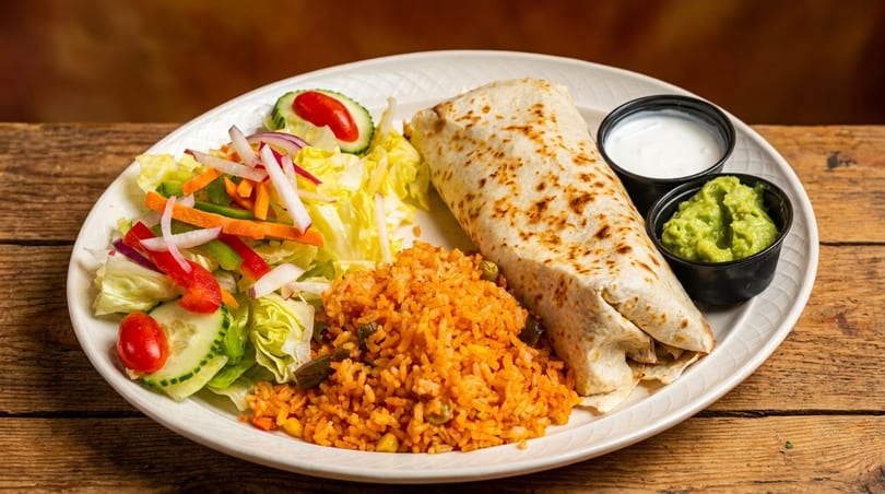 A burrito, Spanish rice, and a side salad with guacamole and sour cream on a white plate.