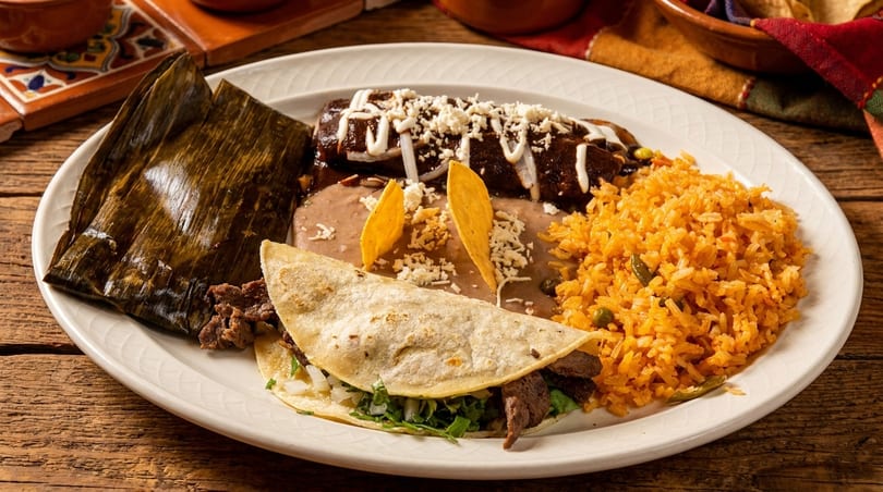 A white plate with a tamale, enchilada, refried beans, rice, and a steak taco on a wooden table.
