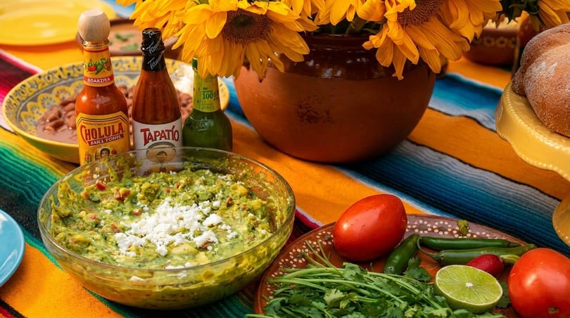 A vibrant Mexican feast with guacamole, hot sauces, fresh vegetables, and sunflowers on a colorful serape.