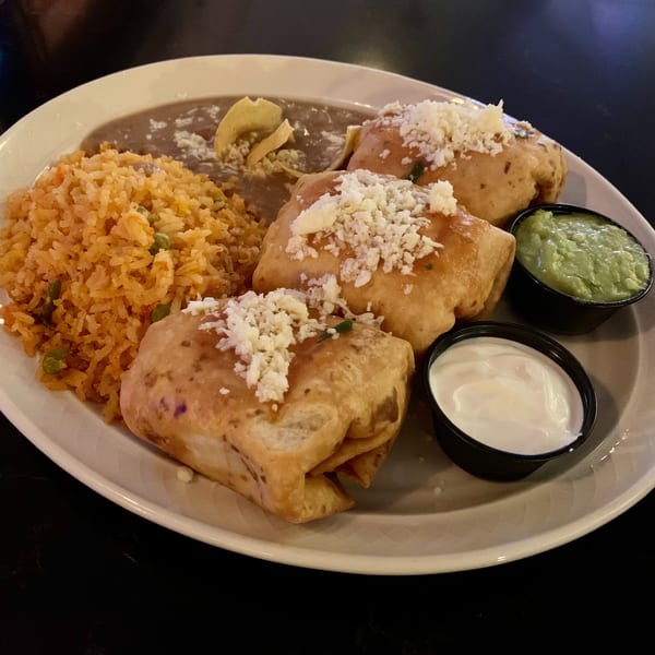 Three golden-brown chimichangas topped with cheese, served with rice, refried beans, guacamole, and sour cream.