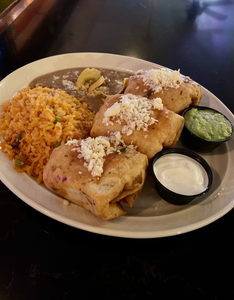 Three golden-brown chimichangas topped with cheese, served with rice, refried beans, guacamole, and sour cream.